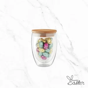 Минималистична чаша от прозрачно стъкло с цилиндрична форма, изпълнена с малки шоколадови яйца в разноцветни станиолени опаковки.